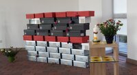 Schuhkartons in den Farben weiß, schwarz und Rot sind zu einer Mauer aufgestapelt. Sie stehen in einem Andachtsraum. Mehrere Schuhkartons in drei Farben bilden eine Mauer.