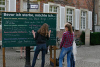 Drei junge Frauen stehen vor einer Tafel, auf der mehrfach der Satzanfang "Bevor ich sterbe, möchte ich ..." geschrieben steht. Man sieht die Frauen von hinten. Eine schreibt etwas auf die Tafel. Drei junge Frauen stehen vor einer Tafel, die in der Öffentlichkeit steht.