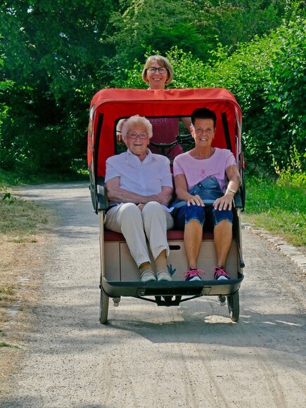 Eine ältere und eine jüngere Frau sitzen in einer Rikscha. Die Rikscha wird von einer Frau gefahren. Die Rikschafahrt findet in einem Park statt. Die Rikscha fährt frontal auf den Betrachter zu. Zwei Frauen sitzen in einer Rikscha